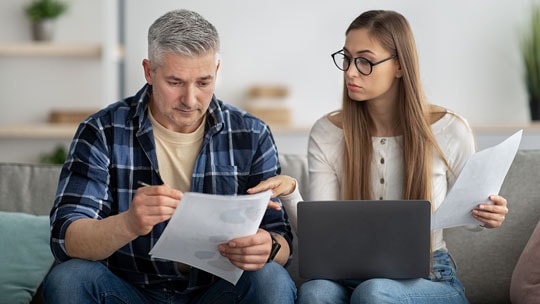 A man and a woman with a laptop on her knees sitting together on a couch reviewing paper statements.